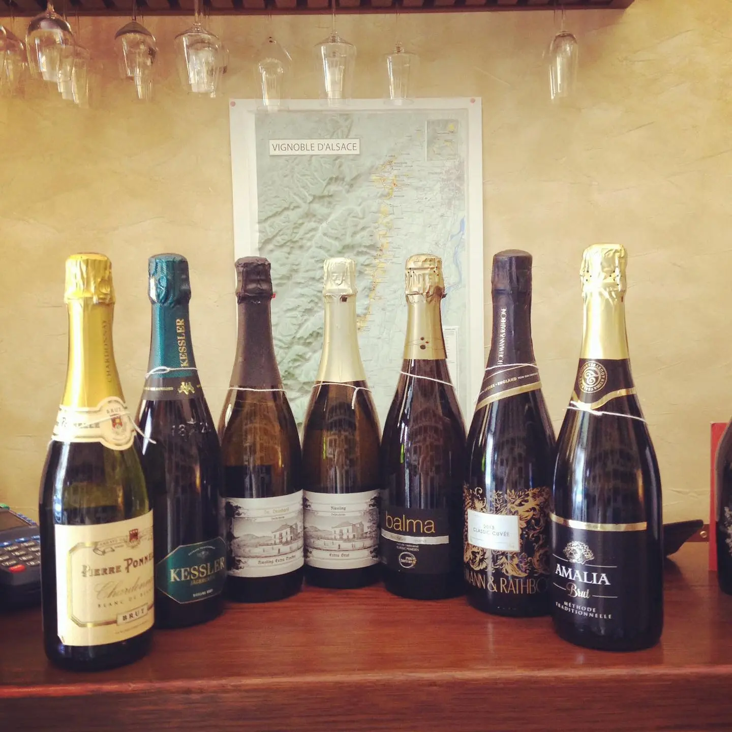 Sette bottiglie di bollicine in fila sul bancone, sotto i calici appesi e davanti alla carta dei vigneti