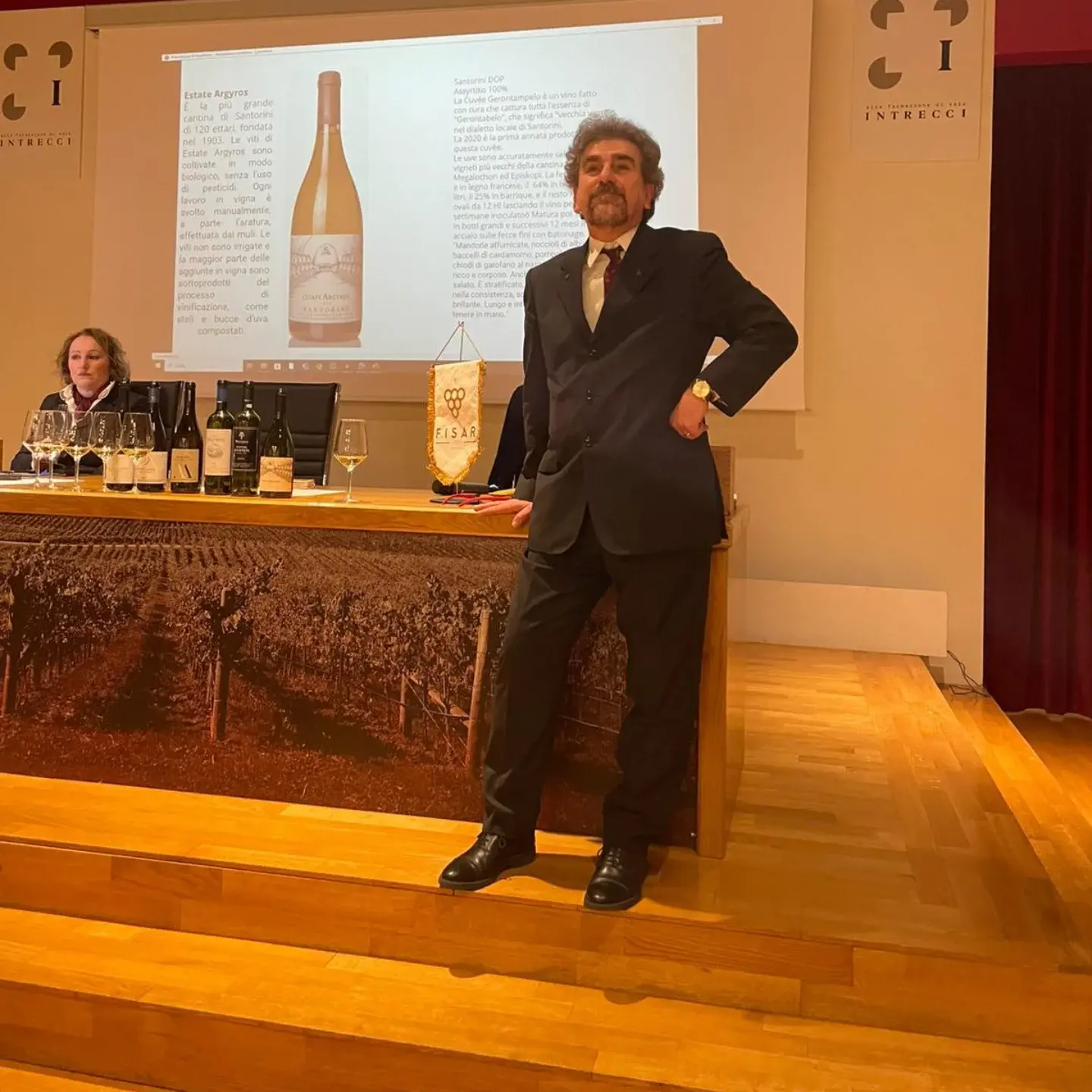 Il Professore in piedi su un palco durante una presentazione, alle spalle la proiezione di una bottiglia