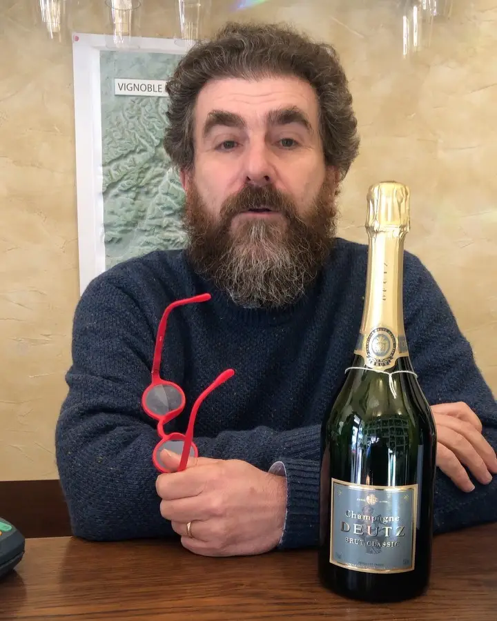 Il Professore con gli occhiali rossi in mano accanto a una bottiglia di Champagne